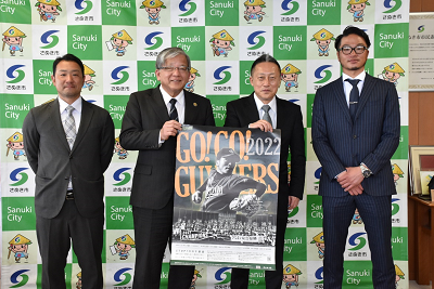 ポスターを手に並んで立っている2人とその左右に立っているスーツ姿の男性2人の写真