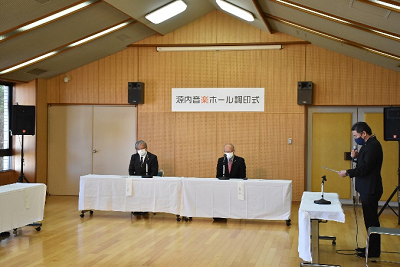 源内音楽ホール調印式と書かれた横断幕の下で司会の男性の話を聞いている男性2人の写真