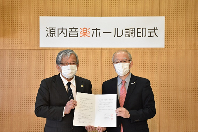 源内音楽ホール調印式と書かれた横断幕の下で一つの書類を両手で持ち正面に掲げている男性2人の写真