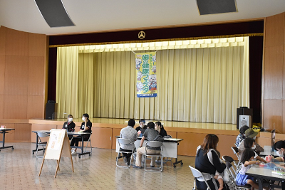 講堂にある複数のテーブルで説明員から説明を受けている人たちの写真