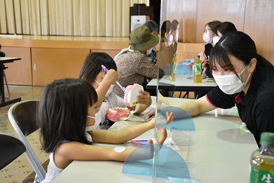 席に着いた大人や子供が対面に座った説明員から歯の模型を使って説明を受けている写真