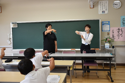 親指と人差し指を立てた右手を下に向ける女性二人とそれを真似る生徒たちの写真