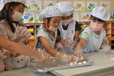 エプロン姿の園児たちがビニール手袋をはめ、丸めた団子をトレーに置いている写真