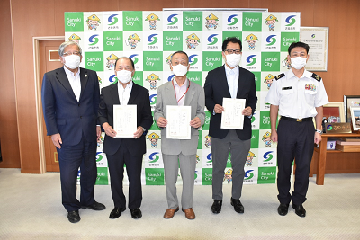 4人のスーツを着た男性と自衛官の男性が並んで立ち、真ん中の男性3人が手に持った紙をカメラに向けている写真