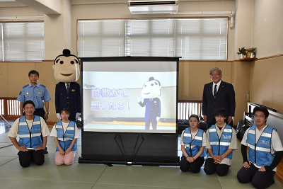 ちょんまげを付けた被り物をした人が写っているスクリーンの周りに座って並んでいる人や立っている人たちがいる写真