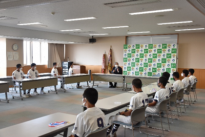 会議室の長机に着席したユニフォーム姿の人たちがスーツ姿の男性の話を聞いている写真