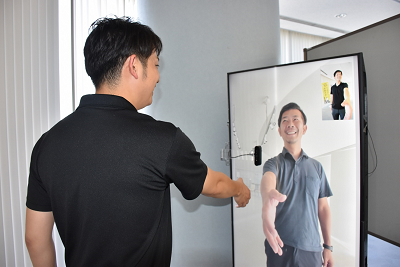 黒のシャツを着た男性がディスプレイに映っている人物と握手をしようとしている写真