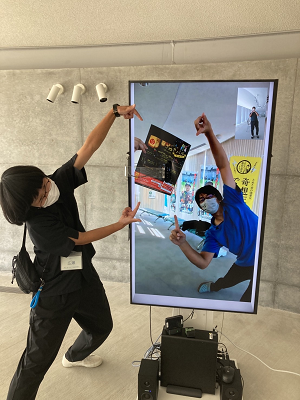 黒のシャツを着ている人がディスプレイに映っている人とポーズを決めている写真