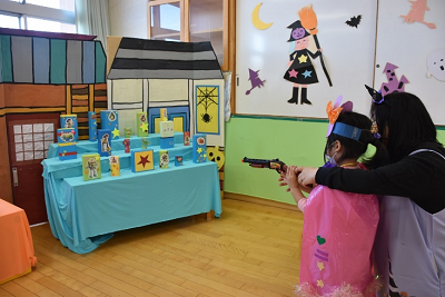 工作で作られた的に向かっておもちゃの銃を構えている園児と一緒に狙いを定めている女性の写真
