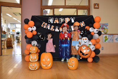黒とオレンジの画用紙で作られたHAPPYHALLOWEENの文字がつるされた黒幕の前でポーズをとる園児とジャックオーランタンの装飾の写真