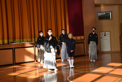 体育館で片手にマイクを持った着物を着た男性の横に立っている金のお面をかぶった児童の写真