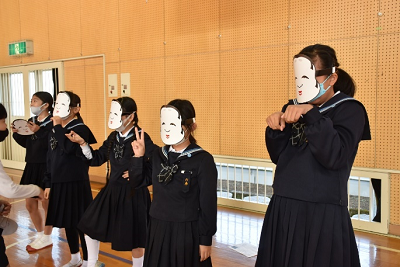 体育館で能面をつけ、各々がポーズをとっている制服姿の女子児童たちの写真