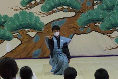 松の絵を背に中腰で閉じた扇子を持った右手を平行に上げている着物を着た人の写真