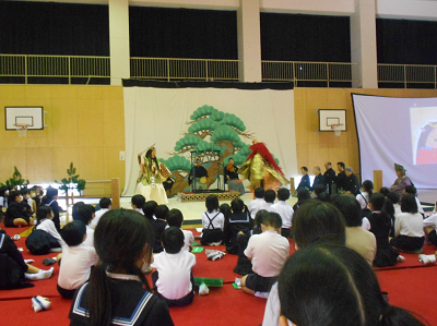 金の着物に鬼のお面をかぶって演技している2名の大人を見学している児童たちの写真