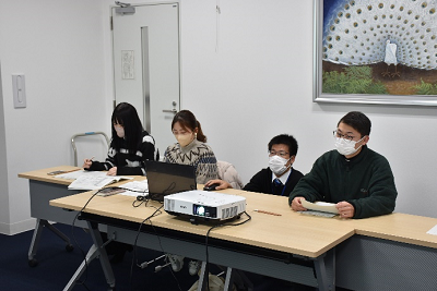 座っている男女3人の間でパソコンのマウスを操作する男性の写真