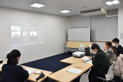 4人の男女が座ってスクリーンに映し出された複数の人と話している写真