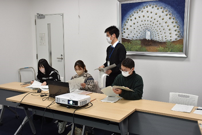 座って手元の資料を見る男女3人の後ろに立つ男性の写真