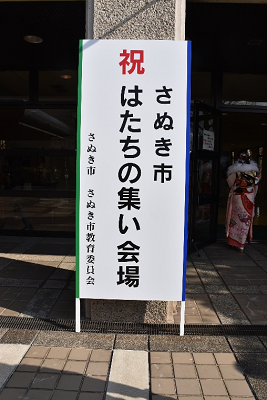 「祝 さぬき市はたちの集い会場」と書かれた看板が柱に立てかけられている写真