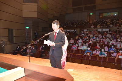 観客席を背にしてステージ上で話すスーツ姿の男性の写真