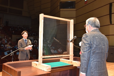 ステージ上でグレーのスーツを着用した男性に向きあって話す男性の写真