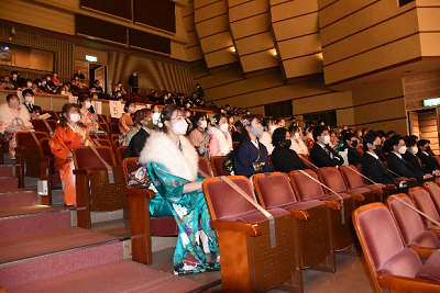 観客席に座り、前方を向く振り袖やスーツを着用した男女の写真