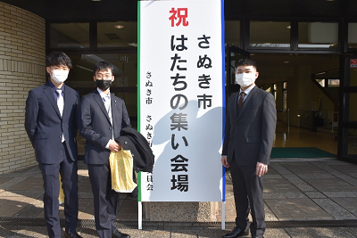 看板の左右に立つスーツ姿の男性3人の写真