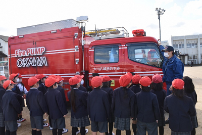 消防車の前に並んで、男性の話を聞く児童たちの写真