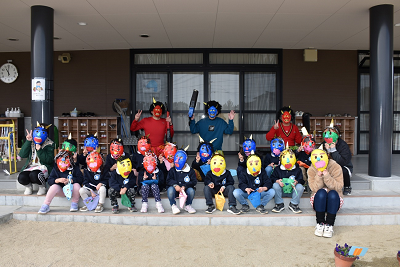 鬼のお面をした園児たちと鬼の仮装をした大人たちが建物の入り口前の階段で並んでポーズをとっている写真