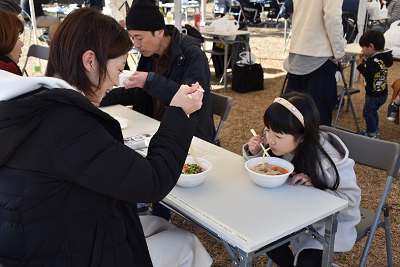 屋外に設置された長机で麺類を食べる親子の写真