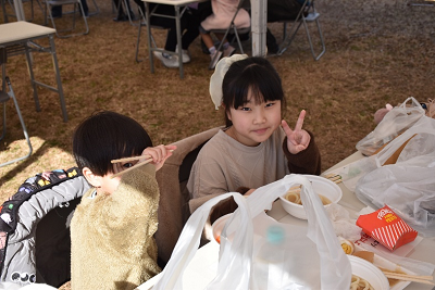 屋外に設置された長机に食べ物を並べ、ピースサインする子供の写真