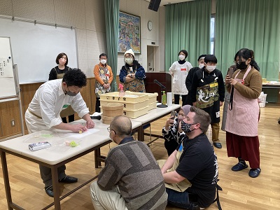 生菓子作りを披露する男性の周りに集まり見学する人達の写真