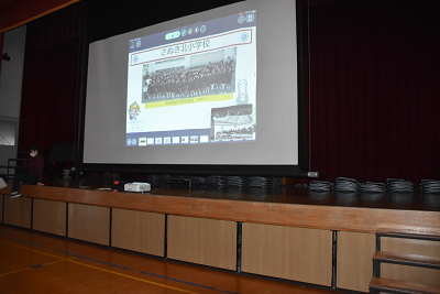 座っている人がいる壇上で展開されているスクリーンに映像が写っている写真