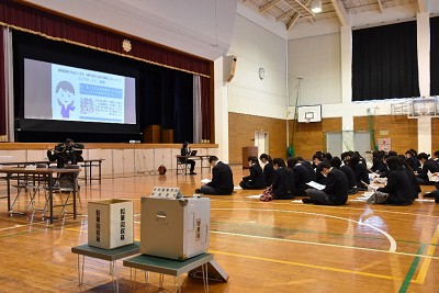 投票箱の置かれた体育館で座り、説明を聞く学生たちの写真
