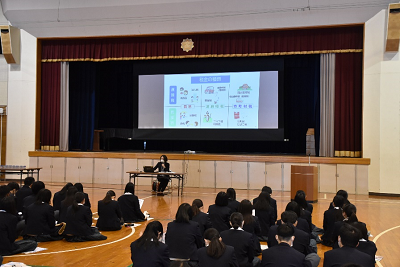 座った生徒たちの前に置かれた机に座って話す女性の写真