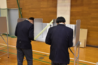 仕切りのついた記載台で用紙に書き込む男子生徒2名の後ろ姿の写真