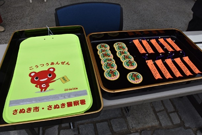 長机に蛍光色で交通安全と書かれたランドセルカバーとワッペン、鈴付きリボンが並べられている写真