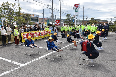 駐車場に並べられた椅子に座っている黄色い帽子をかぶった児童の写真