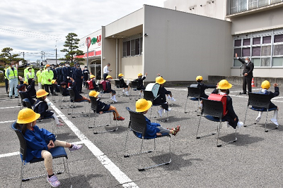 駐車場に並べられた椅子に座っている黄色い帽子をかぶった児童の前で右手を挙げて話をしている男性の写真
