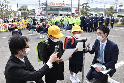 駐車場に設置されたマイクの前で持っている用紙を読み上げている黄色い帽子をかぶった児童の写真
