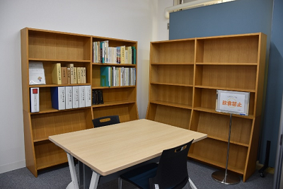 木製の本棚に資料やファイルが並び、その前にテーブルと椅子が置かれた部屋の写真