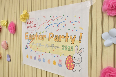 紙で作られた花が飾られた壁に「イースターパーティー2023」と書かれた紙が貼られている写真
