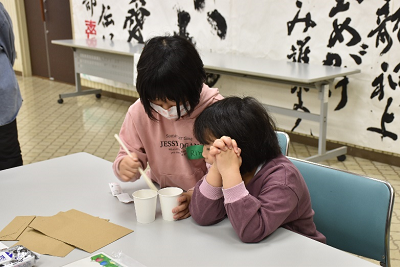 女子児童2人が並んで座り、割りばしと2つの紙コップで遊んでいる写真