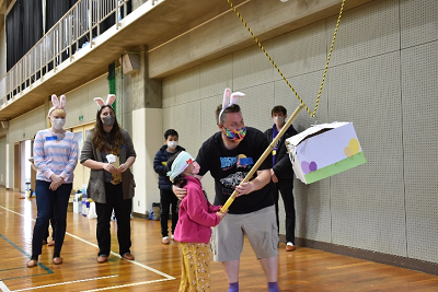 ウサギ耳のカチューシャをつけた男性が目隠しをした女児児童を手伝い、吊り下げられた箱に棒を当てている写真