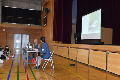 鳥の写真が映し出されたスクリーンの前でパイプ椅子に座り話す男性の写真
