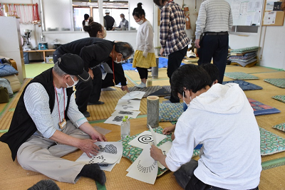 畳みに座り、渦巻きや月などの模様が書かれた複数の紙を並べている写真