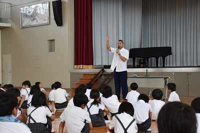 座っている児童の前で話しながら片手をあげる男性の写真