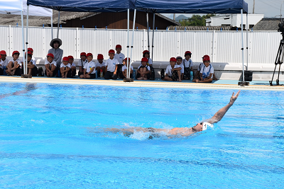 プールサイドで体操服を着た児童たちが座り、プールで背泳ぎする男性を見ている写真