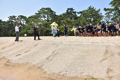 スーツ姿の男性や着ぐるみが見守る中、海に向かって走り出す子供たちの写真
