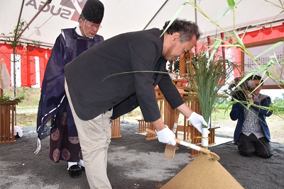 ジャケット姿の男性が盛り砂の頂点に生えた稲を刈っている写真