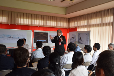 室内で着席した人々の前で話すスーツ姿の男性の写真